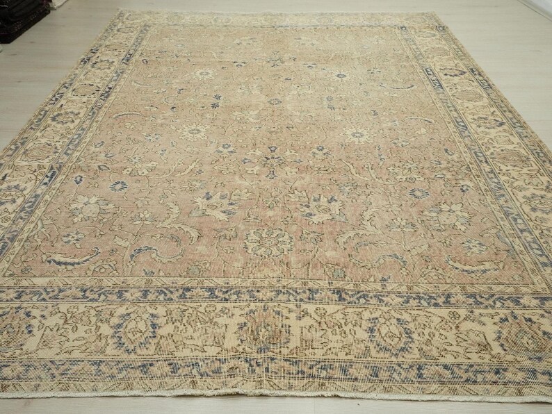 a large, intricately patterned rug with a floral design in shades of beige and blue. The rug is placed on a wooden floor, and the background is a plain white wall.