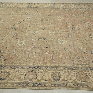 a large, intricately patterned rug with a floral design in shades of beige and blue. The rug is spread out on a wooden floor, and the border is a darker shade of blue.