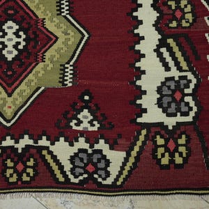 a close-up view of a vibrant, intricately patterned rug with a mix of red, green, and white colors. The rug features a diamond-shaped design in the center, surrounded by floral and geometric patterns in shades of red, green, and white. The rug appears to be a traditional textile, possibly from a Middle Eastern or North African region, with a rich cultural history.