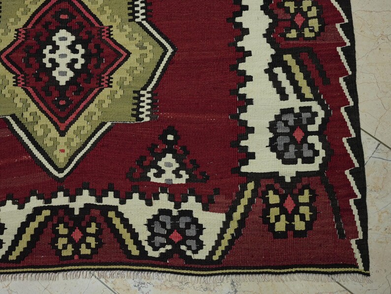 a close-up view of a vibrant, intricately patterned rug with a mix of red, green, and white colors. The rug features a diamond-shaped design in the center, surrounded by floral and geometric patterns in shades of red, green, and white. The rug appears to be a traditional textile, possibly from a Middle Eastern or North African region, with a rich cultural history.
