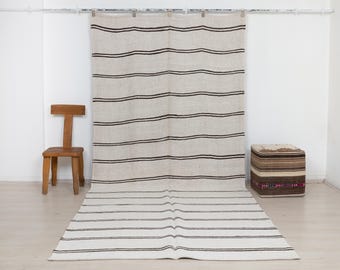 May include: A large, rectangular, off-white textile hanging that extends onto the floor, featuring horizontal dark brown stripes. The fabric has a visible woven texture and is suspended from a metal rod with clips, creating a continuous striped pattern.