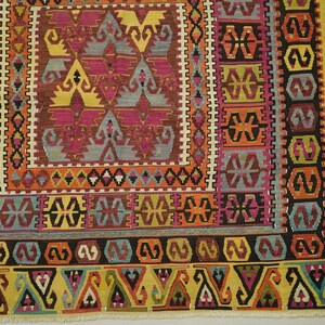 a vibrant and intricate rug with a variety of geometric patterns and designs in a range of colors, including shades of pink, yellow, orange, and green. The rug is framed by a border with a repeating pattern of triangles and diamonds, adding to its decorative appeal. The image is a close-up view of the rug, showcasing its detailed and colorful design.