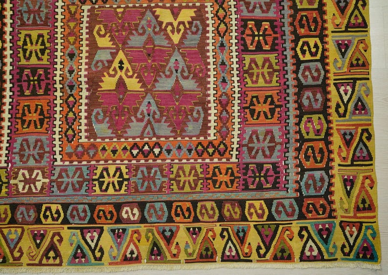 a vibrant and intricate rug with a variety of geometric patterns and designs in a range of colors, including shades of pink, yellow, orange, and green. The rug is framed by a border with a repeating pattern of triangles and diamonds, adding to its decorative appeal. The image is a close-up view of the rug, showcasing its detailed and colorful design.