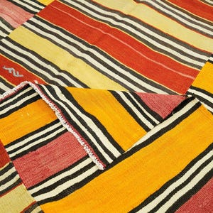 a close-up view of a vibrant, colorful rug with bold stripes in various shades of red, yellow, and black. The rug has a geometric pattern with diagonal stripes and a mix of solid and striped colors, creating a visually striking and eye-catching design.