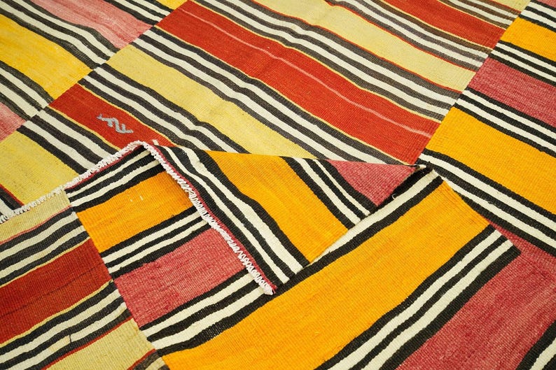 a close-up view of a vibrant, colorful rug with bold stripes in various shades of red, yellow, and black. The rug has a geometric pattern with diagonal stripes and a mix of solid and striped colors, creating a visually striking and eye-catching design.