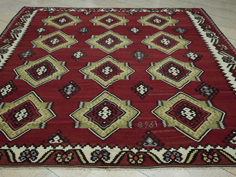 a large, intricately patterned rug with a red background and a border of white and gold designs. The rug features a repeating pattern of diamond shapes in various sizes and colors, creating a visually striking and detailed design.