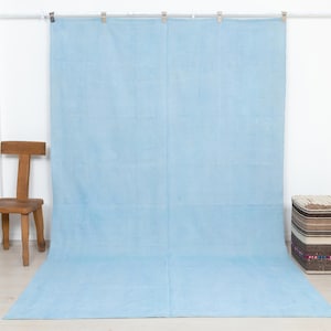 May include: A large, rectangular, light blue fabric backdrop hanging from a metal rod with silver clips. The fabric features a visible vertical seam running down the center and a smooth, matte texture that extends from the wall to the floor.