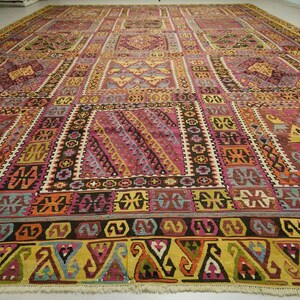 a large, intricately patterned rug with a variety of geometric shapes and designs in a range of colors, including shades of pink, yellow, and orange. The rug is spread out on the floor, covering a significant portion of the space.