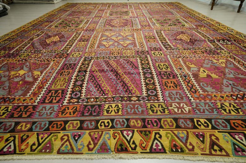 a large, intricately patterned rug with a variety of geometric shapes and designs in a range of colors, including shades of pink, yellow, and orange. The rug is spread out on the floor, covering a significant portion of the space.
