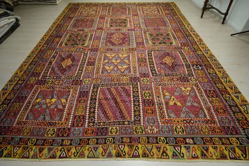 a large, intricately patterned rug with a variety of geometric shapes and designs in shades of pink, yellow, and red. The rug is spread out on a white floor, creating a striking contrast with the colorful patterns.