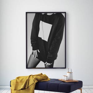 May include: A black and white photograph of a person wearing a black sweatshirt with a white square on the front. The photo is framed in black and hanging on a white wall above a blue upholstered bench with a yellow throw blanket.