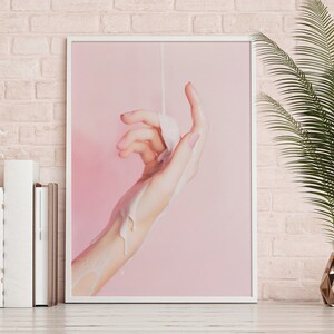 Pu&ograve; includere: Una stampa incorniciata raffigurante una mano con una sostanza bianca versata sopra, su uno sfondo rosa. L'opera d'arte &egrave; esposta su una mensola bianca, con libri e un vaso decorativo con una pianta.