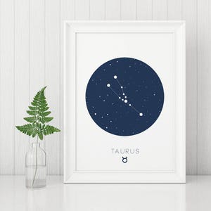 Puede incluir: Una impresión enmarcada en blanco con un círculo azul oscuro que representa la constelación de Tauro con estrellas blancas. El texto "TAURUS" y el símbolo astrológico de Tauro están impresos debajo del círculo.