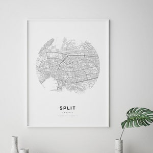 May include: A black and white city map print of Split, Croatia, in a white frame. The map is a circle with the city name and location printed below. The print is on a white wall with two white marble vases and three black vases on a shelf below.