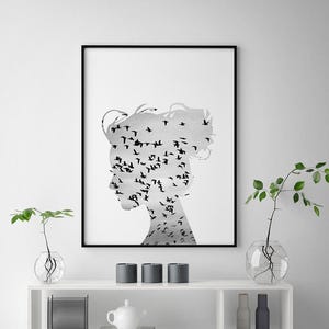 May include: A black and white print of a woman's silhouette with a flock of birds forming her hair and face. The print is framed in a black frame and hanging on a white wall above a white shelf with decorative items.