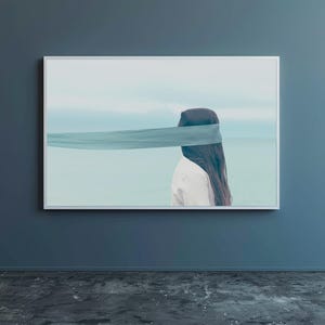 Op de afbeelding: Een ingelijste kunstdruk toont een persoon met lang donker haar en een wit overhemd, met hun ogen bedekt door een turquoise stof. De achtergrond toont een serene zeegezicht met een lichtblauwe lucht en water.