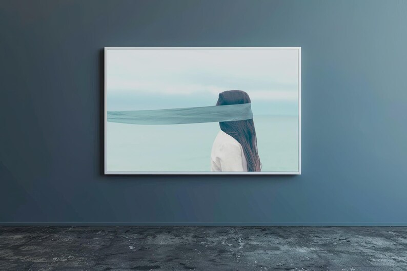 Op de afbeelding: Een ingelijste kunstdruk toont een persoon met lang donker haar en een wit overhemd, met hun ogen bedekt door een turquoise stof. De achtergrond toont een serene zeegezicht met een lichtblauwe lucht en water.