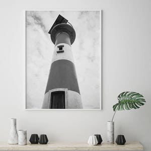 May include: Black and white photograph of a lighthouse against a cloudy sky. The lighthouse is tall and has a white and gray striped exterior. The photo is taken from a low angle, looking up at the lighthouse.