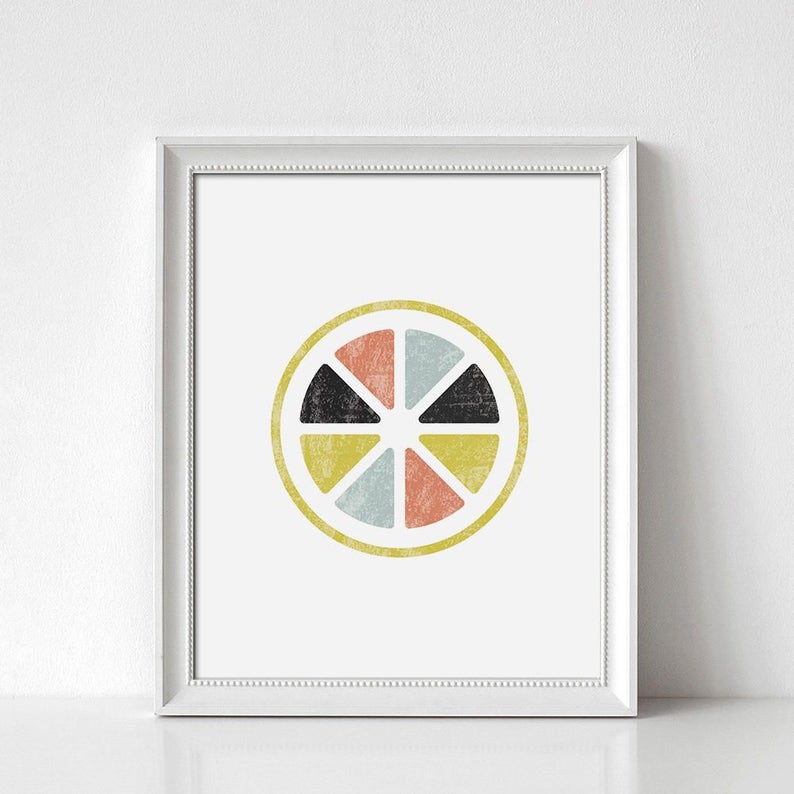 May include: Framed art print featuring a colorful, geometric design of a citrus fruit slice. The artwork includes segments in coral, light blue, yellow, and black, set against a white background. The print is enclosed in a white frame.