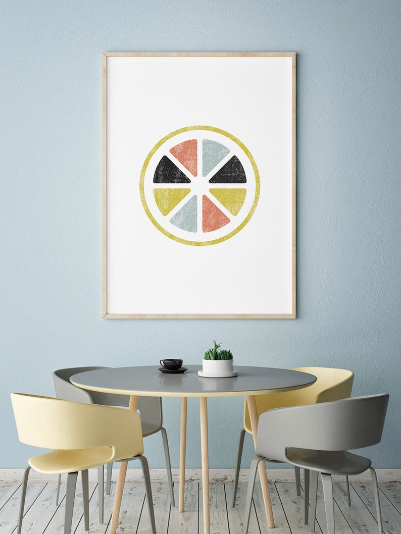 May include: A framed art print featuring a geometric design resembling a citrus fruit slice. The artwork has a yellow outer ring and is divided into eight segments of black, coral, yellow, and blue-green. The print hangs above a gray table and chairs.
