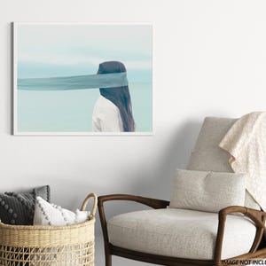 Op de afbeelding: Een ingelijste kunstdruk met een persoon met lang donker haar en een wit overhemd, met een blauwe stof die de ogen bedekt. De achtergrond toont een serene blauwe lucht en water.