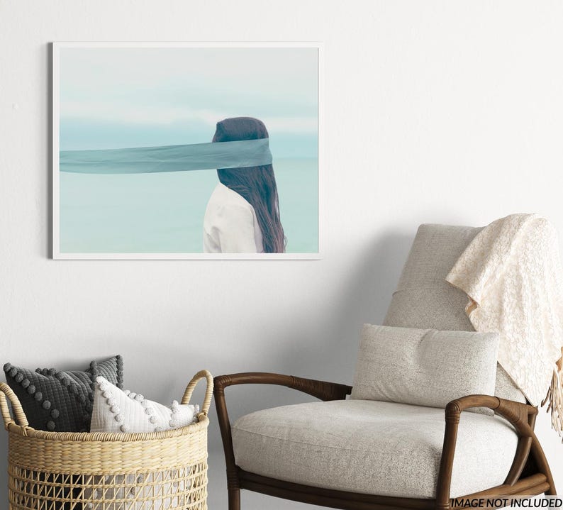 Op de afbeelding: Een ingelijste kunstdruk met een persoon met lang donker haar en een wit overhemd, met een blauwe stof die de ogen bedekt. De achtergrond toont een serene blauwe lucht en water.