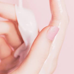 Pu&ograve; includere: Primo piano di una mano con un'unghia dipinta di rosa, che tiene una sostanza bianca e opaca. La pelle appare liscia e leggermente umida, con uno sfondo rosa tenue.
