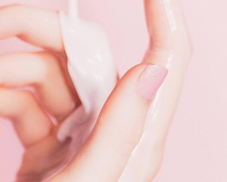 Pu&ograve; includere: Primo piano di una mano con un'unghia dipinta di rosa, che tiene una sostanza bianca e opaca. La pelle appare liscia e leggermente umida, con uno sfondo rosa tenue.