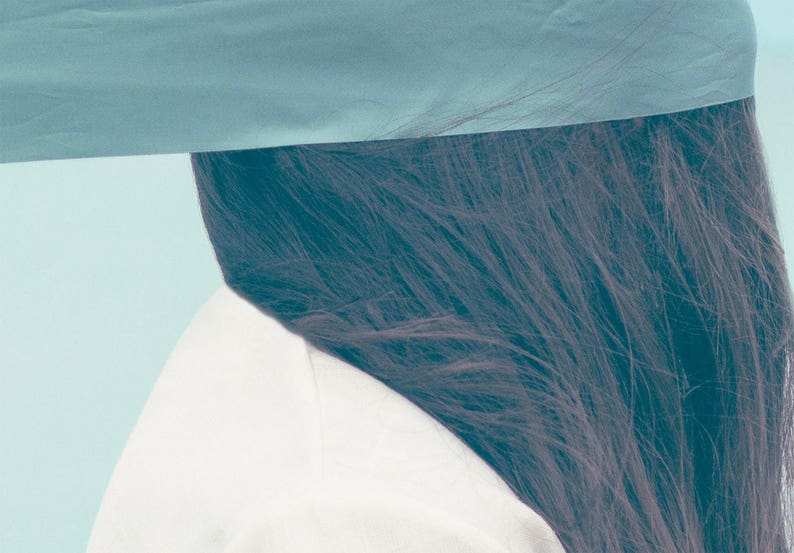 Op de afbeelding: Een artistieke afbeelding met donker haar, een wit kledingstuk en een blauwgroene stof. De stof is over de bovenkant van het frame gedrapeerd en verbergt gedeeltelijk het haar en het witte kledingstuk. De achtergrond is lichtblauwgroen.