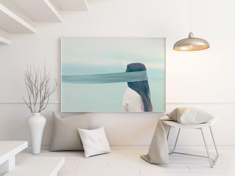 Op de afbeelding: Een ingelijste kunstdruk toont een persoon met lang donker haar en een turquoise blinddoek tegen een serene, lichtblauwe achtergrond. Het kunstwerk hangt boven een witte bank met kussens en een decoratieve vaas met takken.
