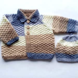 Op de afbeelding: Een gebreide baby set in crème, bruin en blauw. De set bevat een trui met kraag en houten knopen en een bijpassende muts. De trui en muts hebben een textuur steekpatroon.