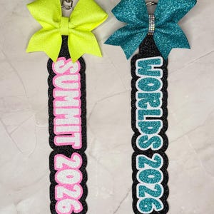 May include: Two keychains with glitter bows. One is neon yellow, the other teal. Both have a black base with the words "SUMMIT 2026" and "WORLDS 2026" in pink and teal glitter, respectively. Each keychain has a silver clasp.