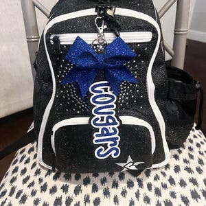 May include: Black and white glitter cheerleading backpack with a blue bow and a white patch that says "Superstars".