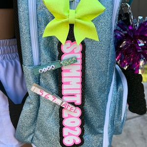 May include: A teal backpack with a neon yellow bow and a pink and black Summit 2026 tag. Two clothespins are attached, one with the word "DIVA" and the other with the numbers "567...ATEL". A purple pom-pom is visible.