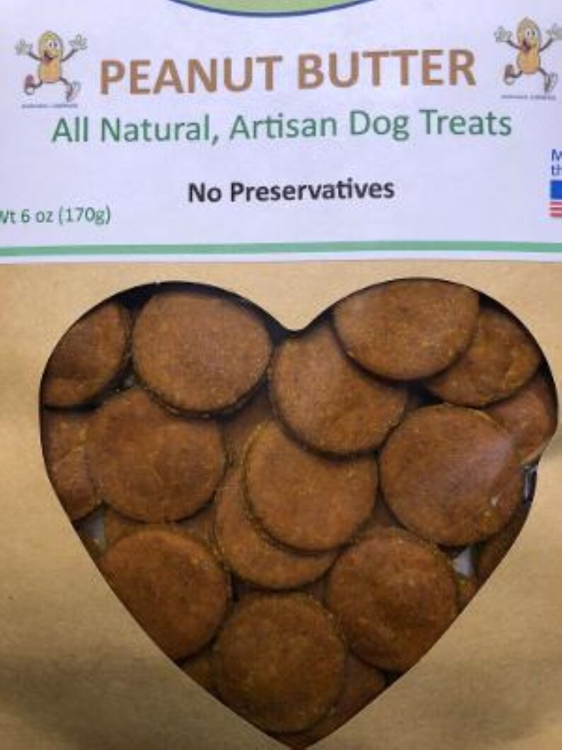 May include: A brown paper bag with a heart-shaped window showing a pile of peanut butter dog treats. The bag has the text "PEANUT BUTTER" in green letters and "All Natural, Artisan Dog Treats" in black letters. The bag also has the text "No Preservatives" and "Wt 6 oz (170g)".