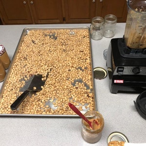 May include: A metal baking sheet filled with roasted peanuts, a spatula, and a jar of peanut butter. There is a blender with peanut butter residue on the side and a jar of peanut butter on the counter.