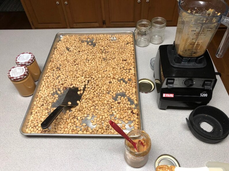 May include: A metal baking sheet filled with roasted peanuts, a spatula, and a jar of peanut butter. There is a blender with peanut butter residue on the side and a jar of peanut butter on the counter.