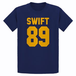 May include: Navy blue t-shirt with the word "SWIFT" in gold block letters above the number "89" in gold. The shirt is a classic crew neck style.