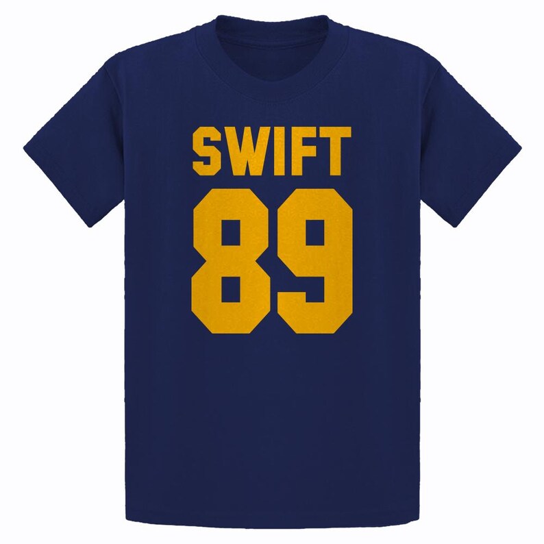 May include: Navy blue t-shirt with the word "SWIFT" in gold block letters above the number "89" in gold. The shirt is a classic crew neck style.