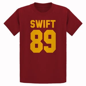 May include: Maroon t-shirt with the word "SWIFT" and the number "89" in gold block letters. The shirt has short sleeves and a crew neck. The text and numbers are centered on the front of the shirt.