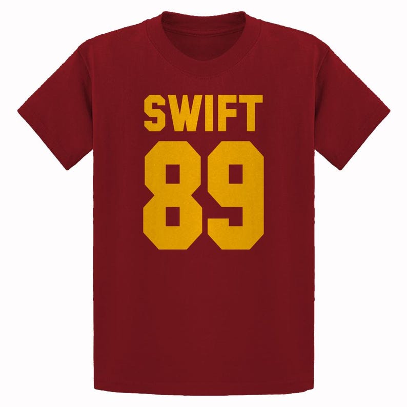 May include: Maroon t-shirt with the word "SWIFT" and the number "89" in gold block letters. The shirt has short sleeves and a crew neck. The text and numbers are centered on the front of the shirt.