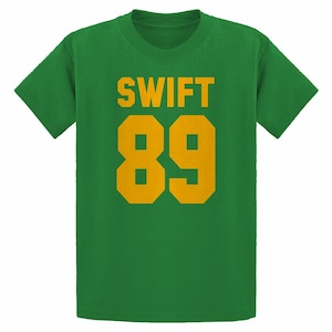 May include: Green t-shirt with the word "SWIFT" in gold block letters above the number "89" in gold. The shirt is a solid green color and has short sleeves.