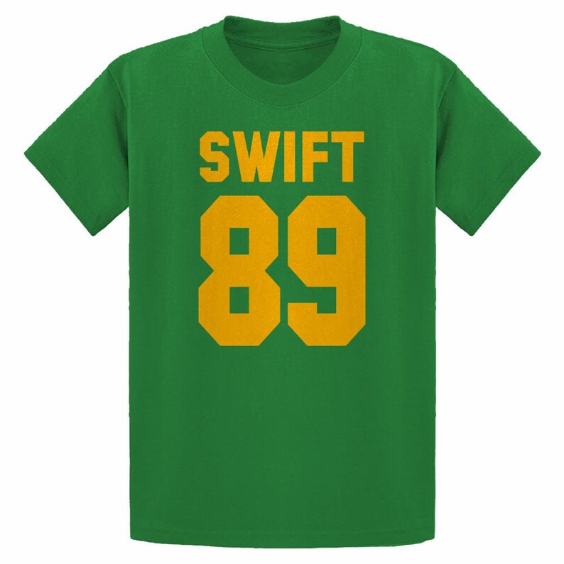May include: Green t-shirt with the word "SWIFT" in gold block letters above the number "89" in gold. The shirt is a solid green color and has short sleeves.