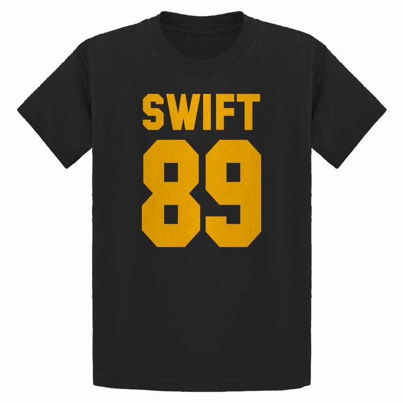 May include: Black t-shirt with the word "SWIFT" and the number "89" printed in gold. The text and numbers are in a bold, blocky font. The shirt has short sleeves and a crew neck.