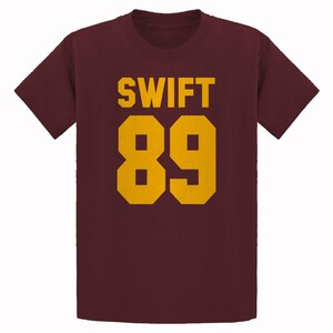 May include: Maroon t-shirt with the word "SWIFT" and the number "89" printed in gold. The text and numbers are in a bold, blocky font. The shirt has short sleeves and a crew neck.
