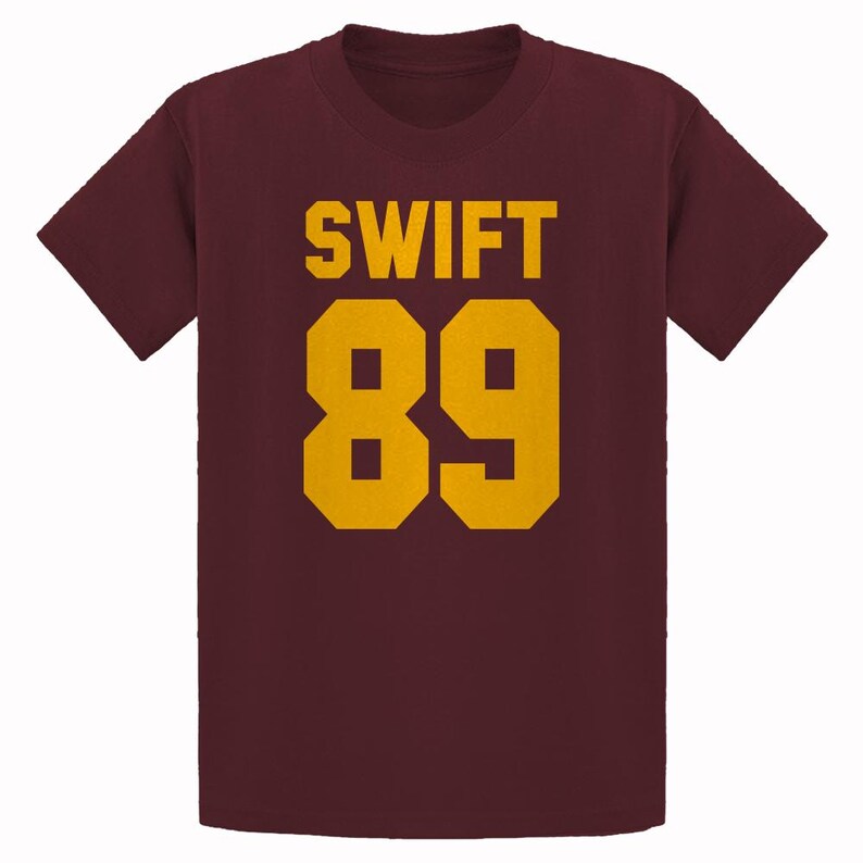 May include: Maroon t-shirt with the word "SWIFT" and the number "89" printed in gold. The text and numbers are in a bold, blocky font. The shirt has short sleeves and a crew neck.