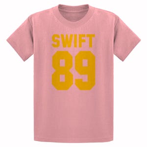 May include: A light pink t-shirt with the word "SWIFT" in yellow block letters above the number "89" in yellow block letters. The shirt has short sleeves and a crew neck.
