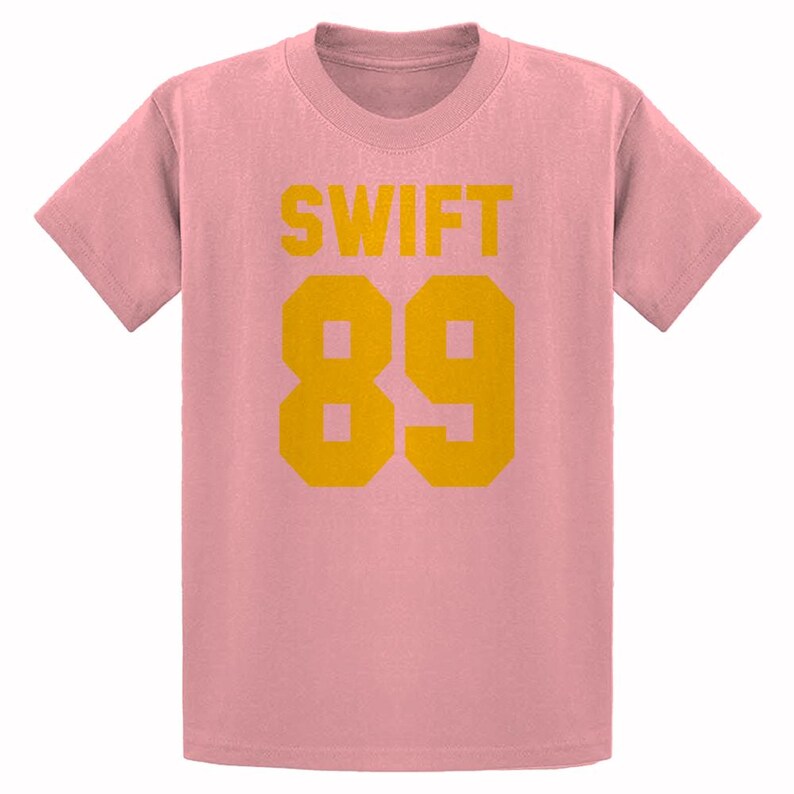 May include: A light pink t-shirt with the word "SWIFT" in yellow block letters above the number "89" in yellow block letters. The shirt has short sleeves and a crew neck.