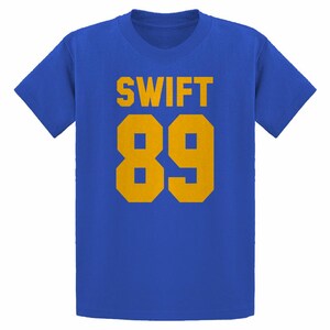 May include: Royal blue t-shirt with the word "SWIFT" in gold block letters above the number "89" in gold. The shirt is a crew neck style and has short sleeves. The design is centered on the front of the shirt.