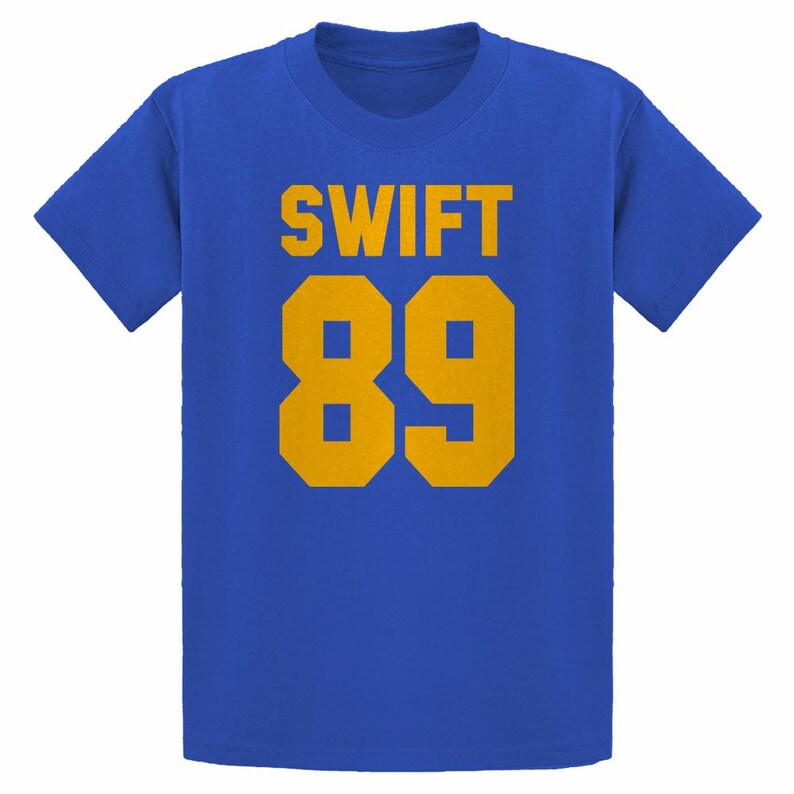 May include: Royal blue t-shirt with the word "SWIFT" in gold block letters above the number "89" in gold. The shirt is a crew neck style and has short sleeves. The design is centered on the front of the shirt.
